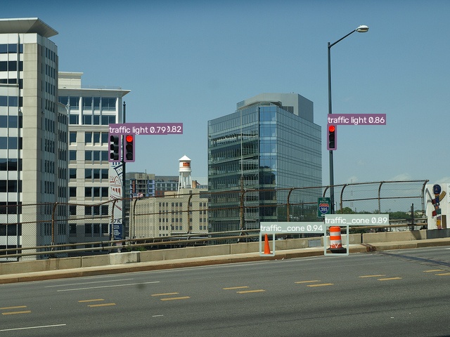 A highway overpass shot from across the road: two traffic cones on the far shoulder detected at 0.94 and 0.89, plus three traffic lights against a city skyline